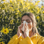 Woman blowing her nose with a tissue near yellow flowers, a sign of fall allergies and sinus congestion